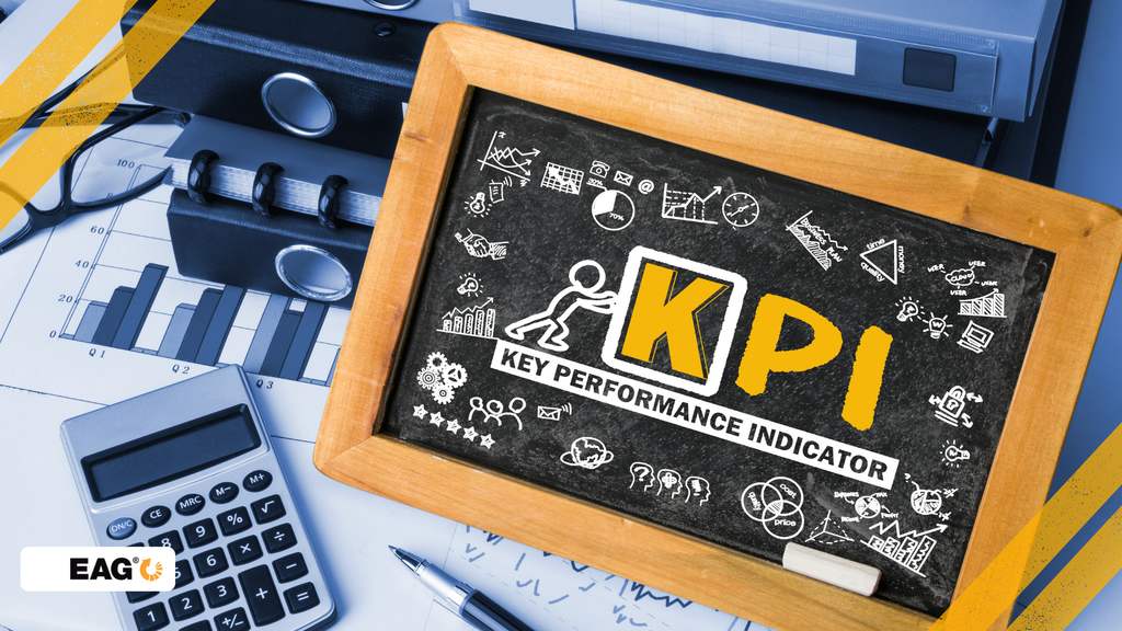Sigla KPI desenhada num quadro com elementos que simbolizam tipos de indicadores de desempenho de uma empresa. Sobre a mesa, uma calculadora e óculos sinalizando ambiente de trabalho